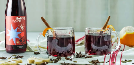 750ml bottle of Holiday Spirit is to the left of two mugs of mulled wine that are garnished with cinnamon sticks and orange peels