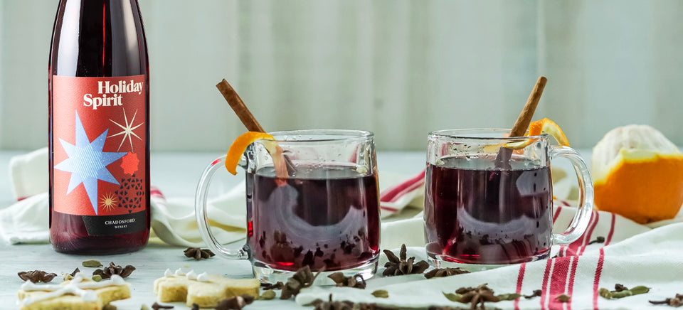 750ml bottle of Holiday Spirit is to the left of two mugs of mulled wine that are garnished with cinnamon sticks and orange peels