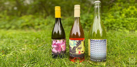 Three 750ml bottles of Sparkling White, '19 Dry Rosé and '19 Vignoles are placed on a grassy landscape. 