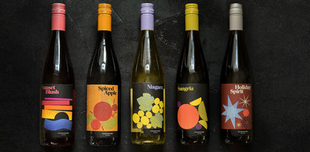 Five sweet wine bottles including Sunset Blush, Spiced Apple, Niagara, Sangria and Holiday Spirit lay next to each other on a black slate background. 